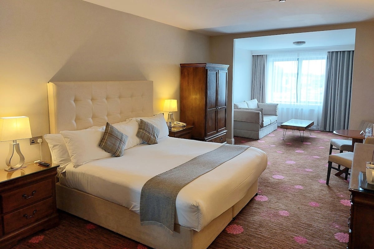 A hotel room with a large bed, a wooden wardrobe, a seating area with a sofa and armchairs, and a small table with a lamp.