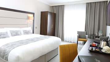 A hotel room with a large bed, a desk with a tray of breakfast items, a chair, a wardrobe, and a large window with curtains.