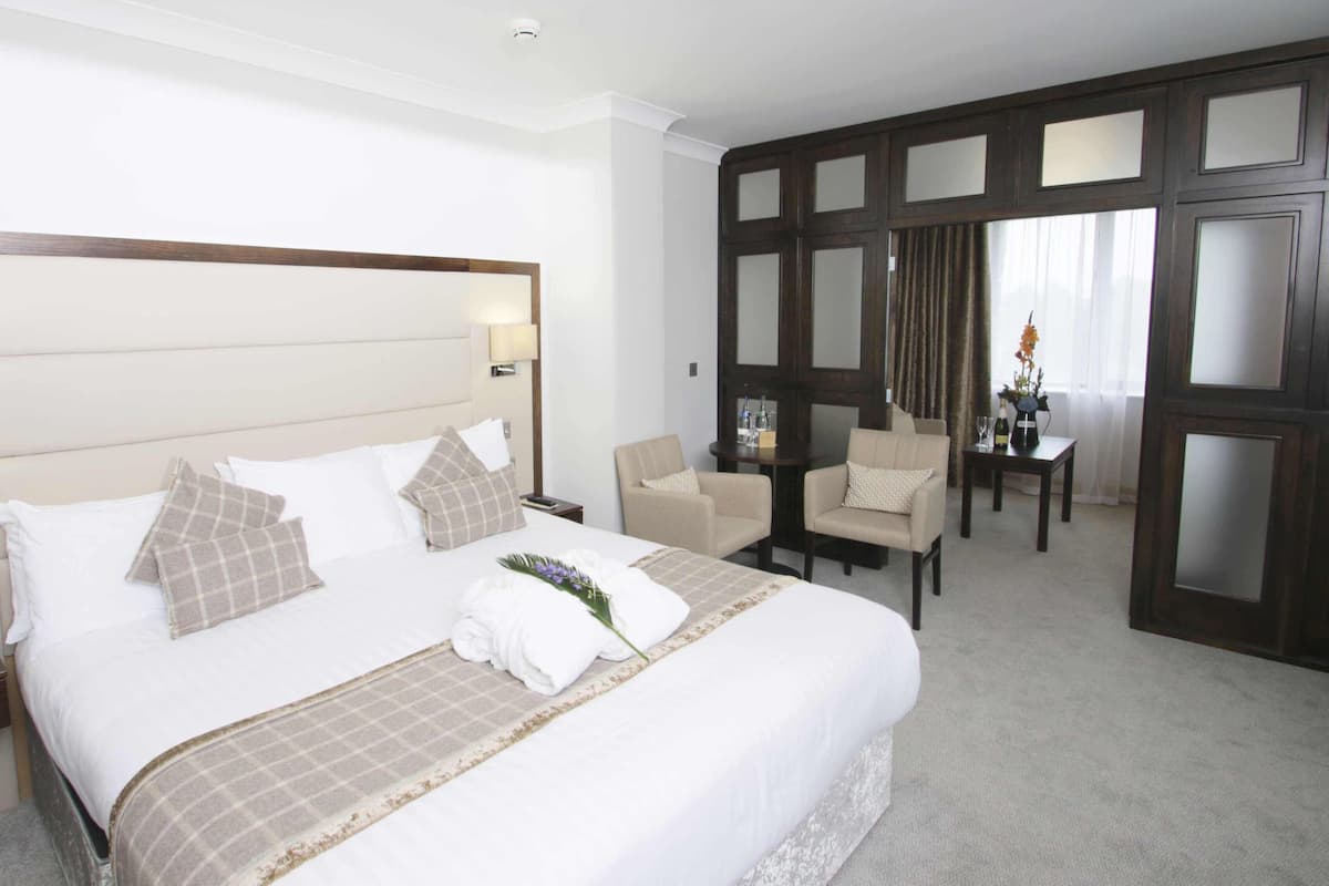 A hotel room with a large bed, two bedside tables, a chair, a small table with a vase, and a dark wooden partition with glass panels.