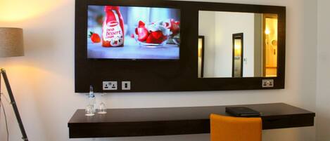A hotel room with a TV displaying a dessert advertisement, a desk with a laptop, a chair, and a trash can.