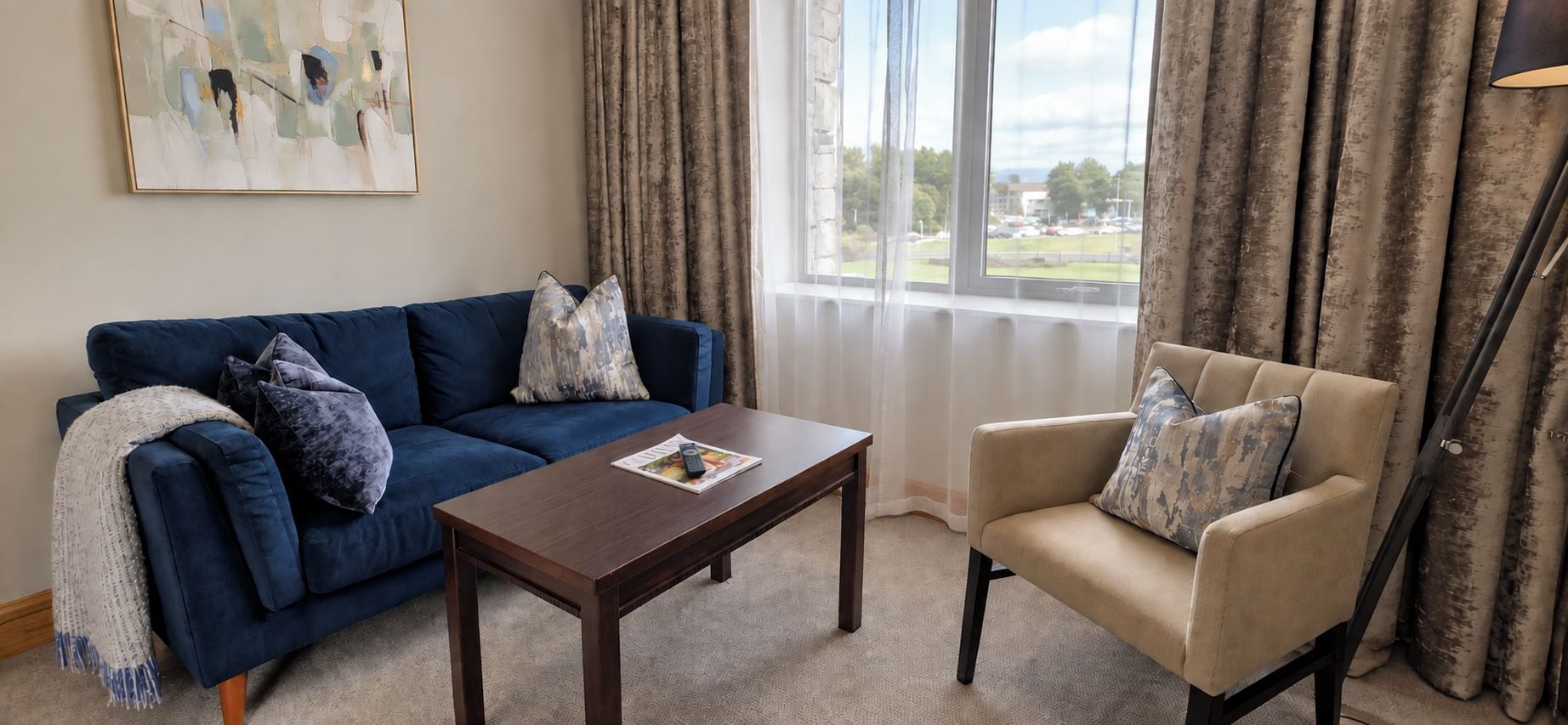 A living room with a blue sofa, a beige armchair, a wooden coffee table, and a view through a window with curtains.