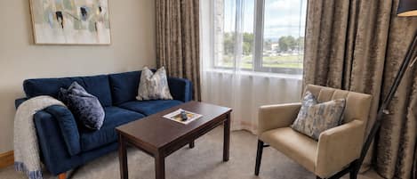 A living room with a blue sofa, a beige armchair, a wooden coffee table, and a view through a window with curtains.
