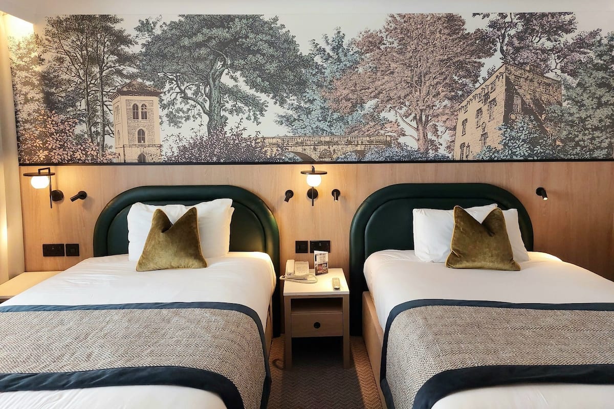 A hotel room with two beds, a nightstand with a phone and remote, and a wall tapestry depicting trees and buildings.