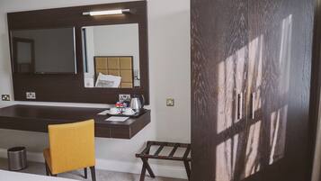 Quarto de hotel com uma cama, uma escrivaninha com cadeira, um guarda-roupa, um espelho e uma pequena mesa com chaleira e chávenas.