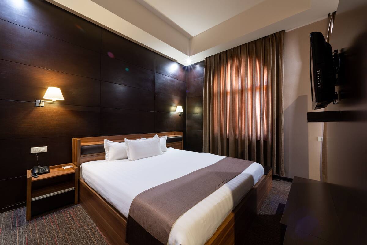 Une chambre d’hôtel avec un grand lit, une tête de lit en bois, deux tables de chevet avec des lampes, un téléviseur à écran plat fixé au mur et une fenêtre avec des rideaux.