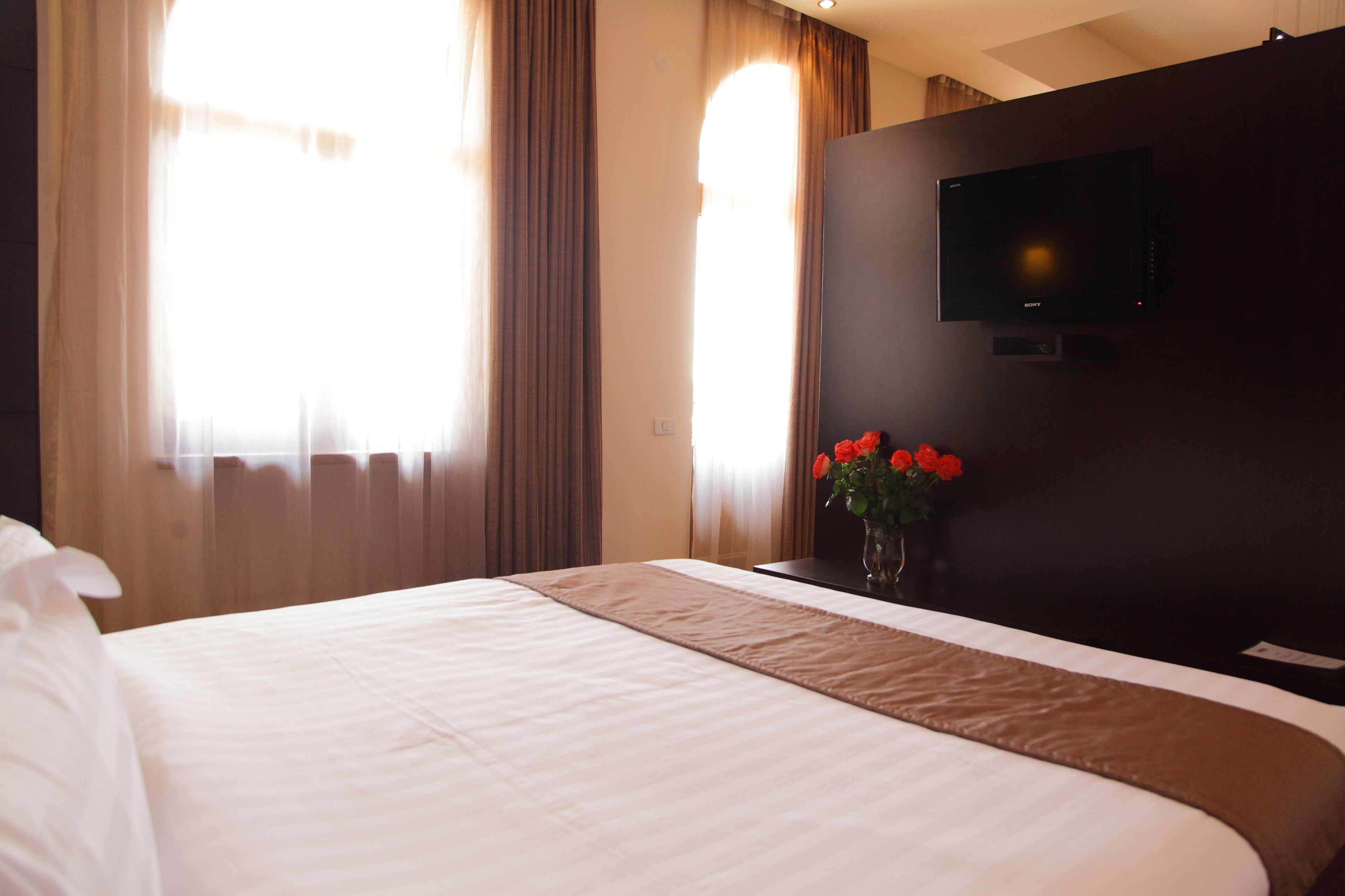 Quarto de hotel com uma cama grande, uma televisão de tela plana na parede e um vaso com flores vermelhas na mesa de cabeceira.