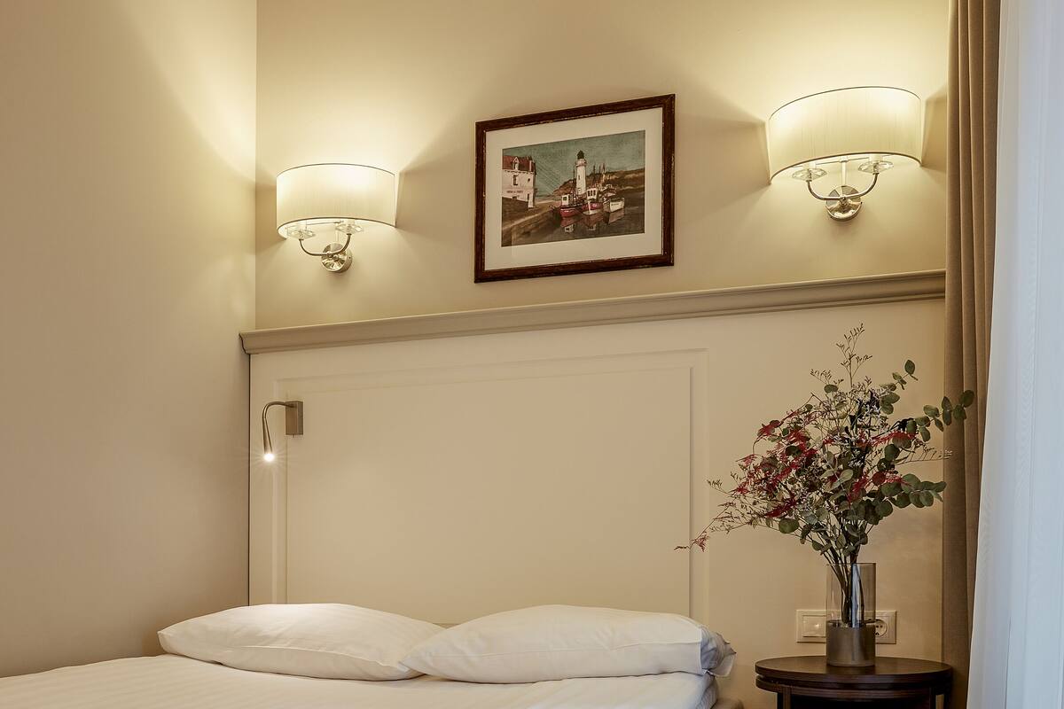 A hotel room with a neatly made bed, a small bedside table with a vase of flowers, and two wall-mounted lamps.