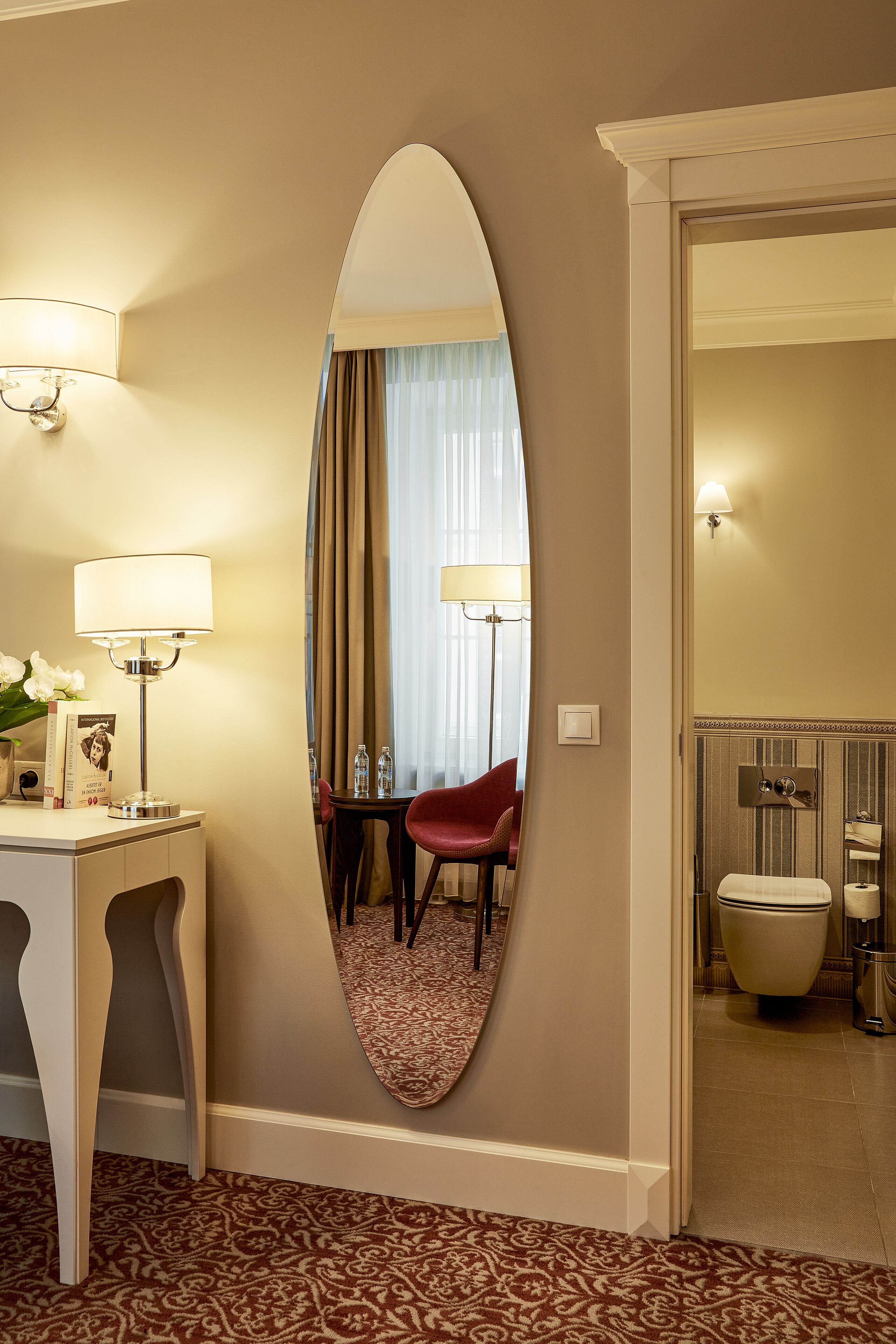 Habitación de hotel con un espejo ovalado grande, un escritorio con lámpara, una silla y una cama con inodoro y bidé.