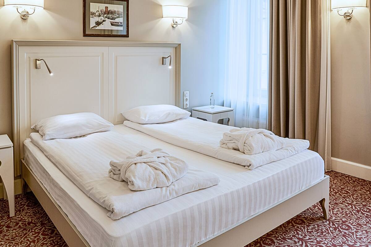 A hotel room with a large bed, two pillows, a bedside table with a bottle, and a framed picture on the wall.