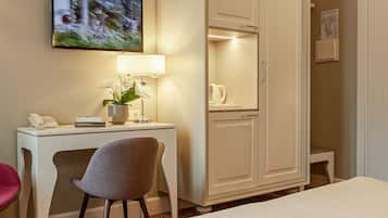 A hotel room with a bed, a desk with a lamp, a chair, a wardrobe, and a painting on the wall.