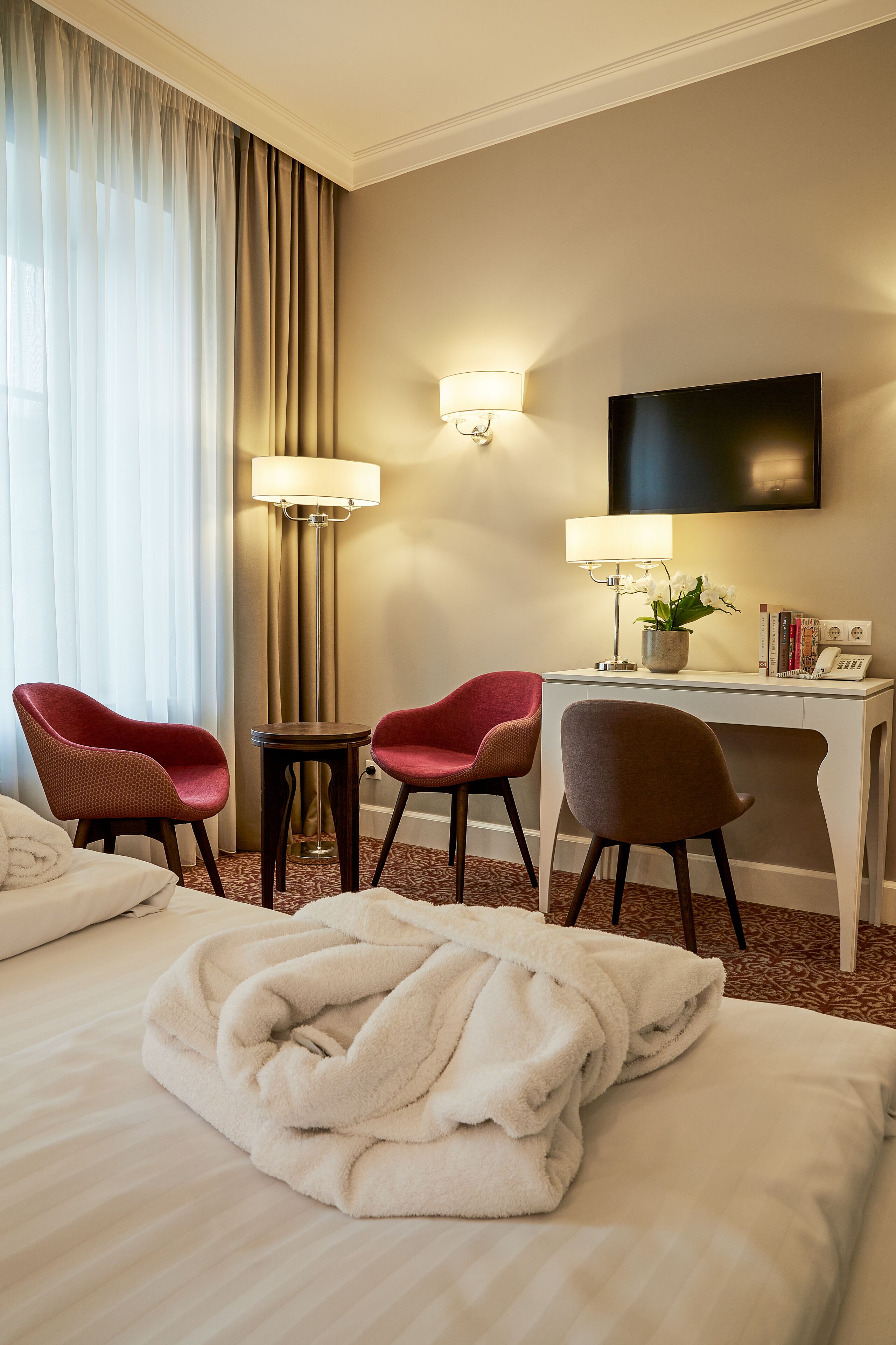 A hotel room with a bed, two chairs, a desk, a lamp, a TV, and a telephone.