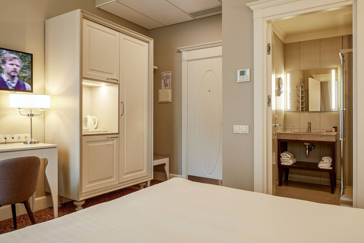 A hotel room with a bed, a desk with a lamp, a wardrobe, a bathroom with a sink and mirror, and a television on the wall.