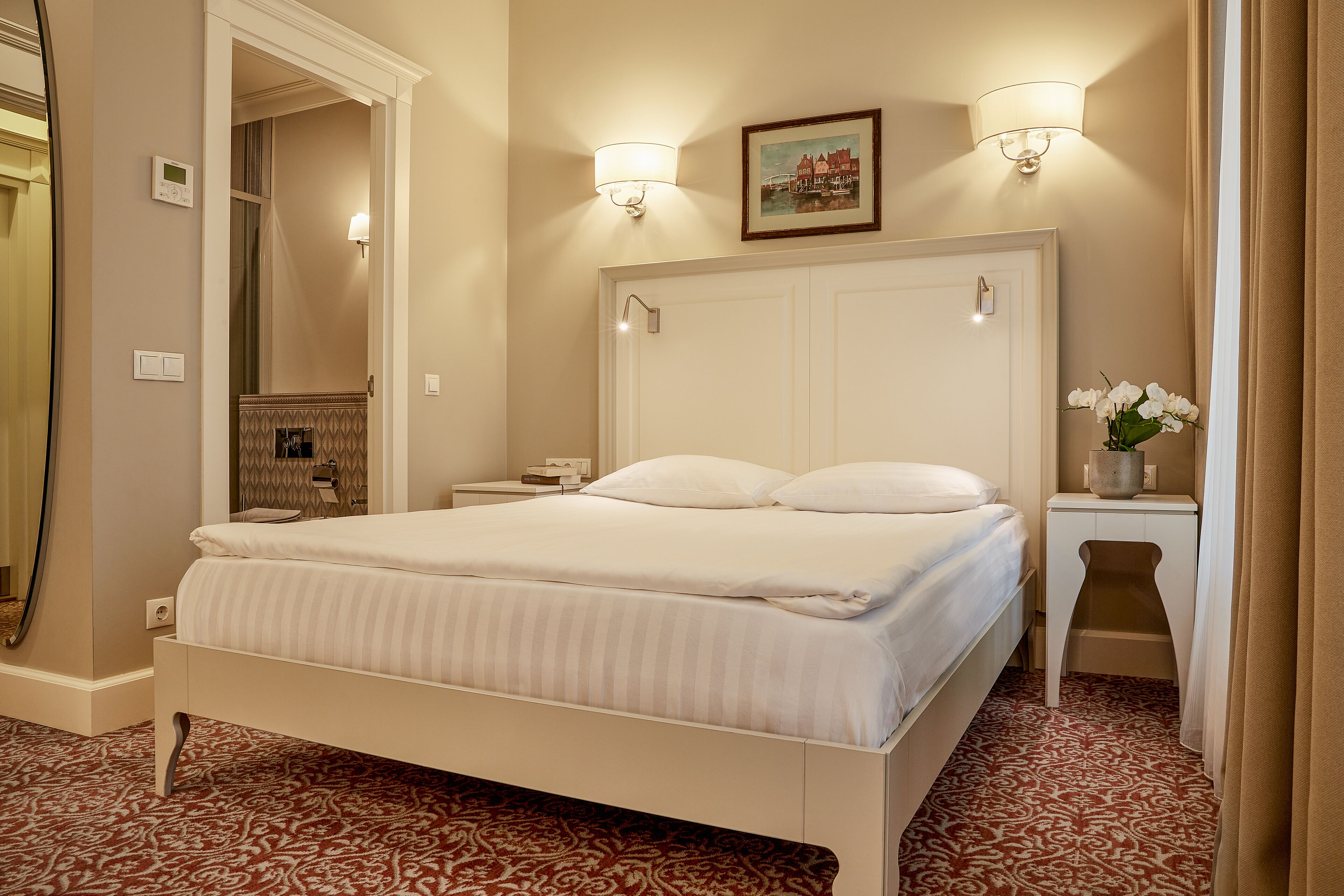 Een net opgemaakt bed in een hotelkamer met een kamerbreed tapijt, een nachtkastje met een lamp en een ingelijst schilderij aan de muur.