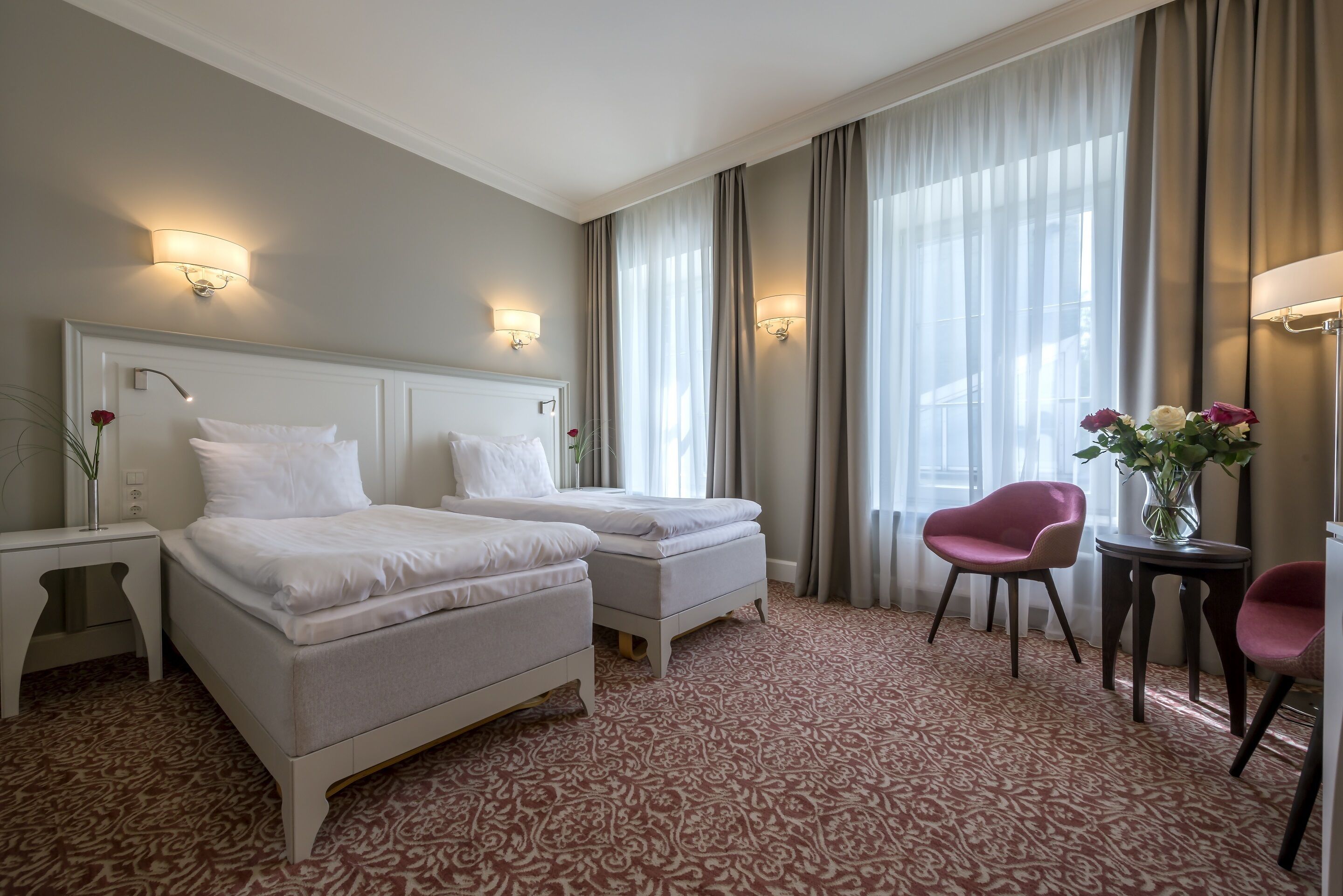 Habitación de hotel con dos camas, una zona de estar con una silla y una mesa, y un jarrón con flores.