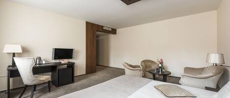 A hotel room with a bed, desk, two chairs, a TV, and a small table with a lamp and flowers.