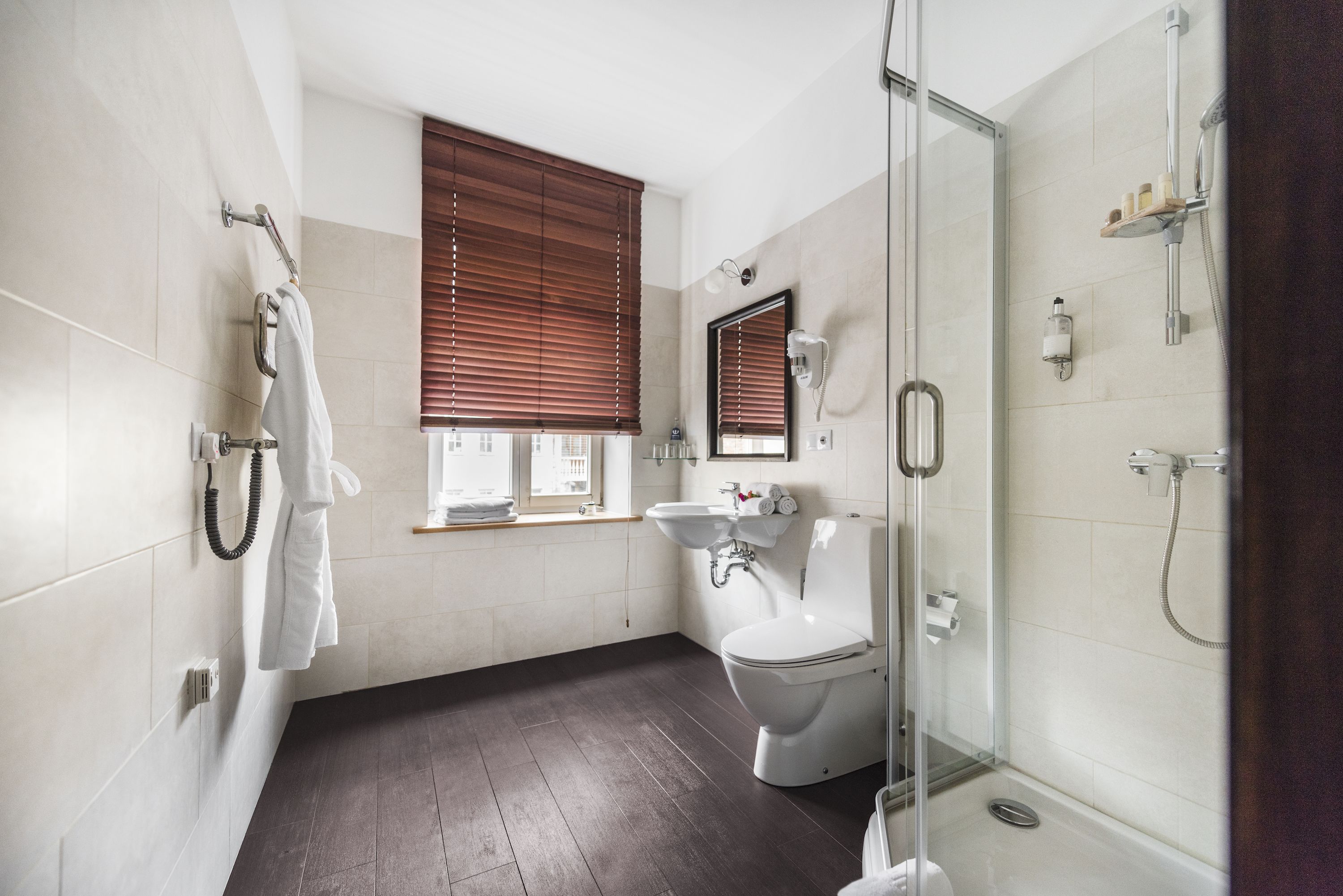 Ein modernes Badezimmer mit Toilette, Waschbecken und Dusche. An den Fenstern befinden sich Holzjalousien, und ein Handtuchhalter ist zu sehen.