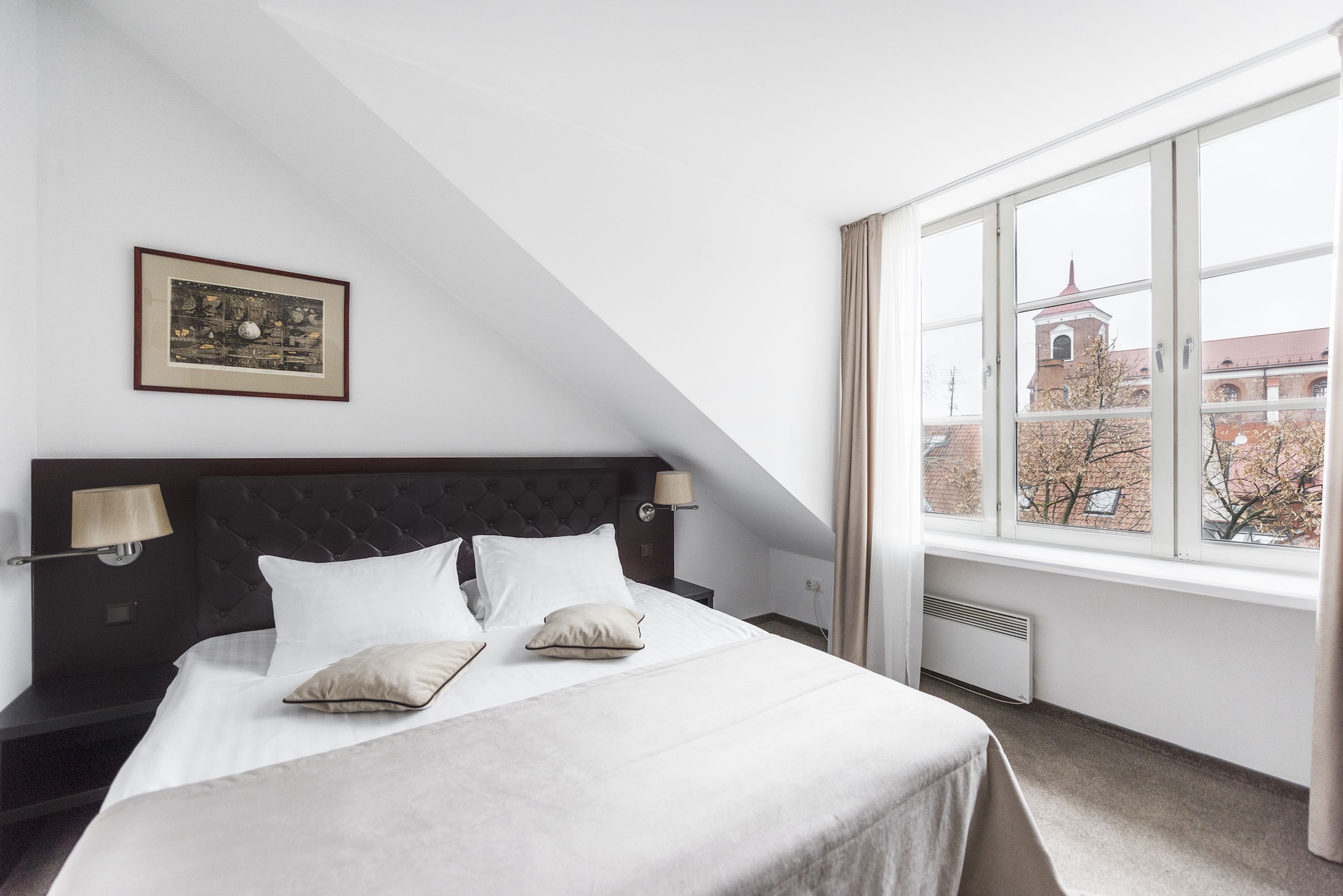 Et pænt indrettet hotelværelse med en stor seng, et billede i ramme på væggen og et vindue med udsigt til en bygning med et tårn.