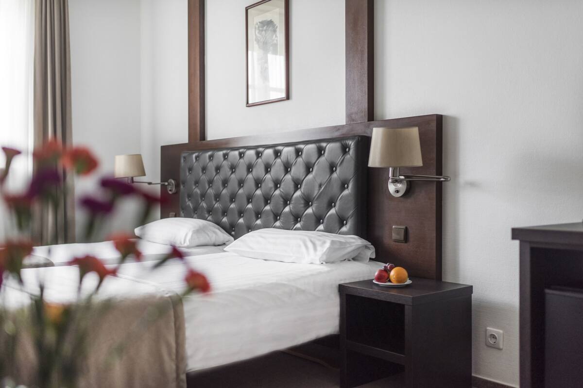 Un lit bien fait, avec du linge de lit blanc et une tête de lit capitonnée, une table de chevet avec une lampe et un tableau encadré au mur.