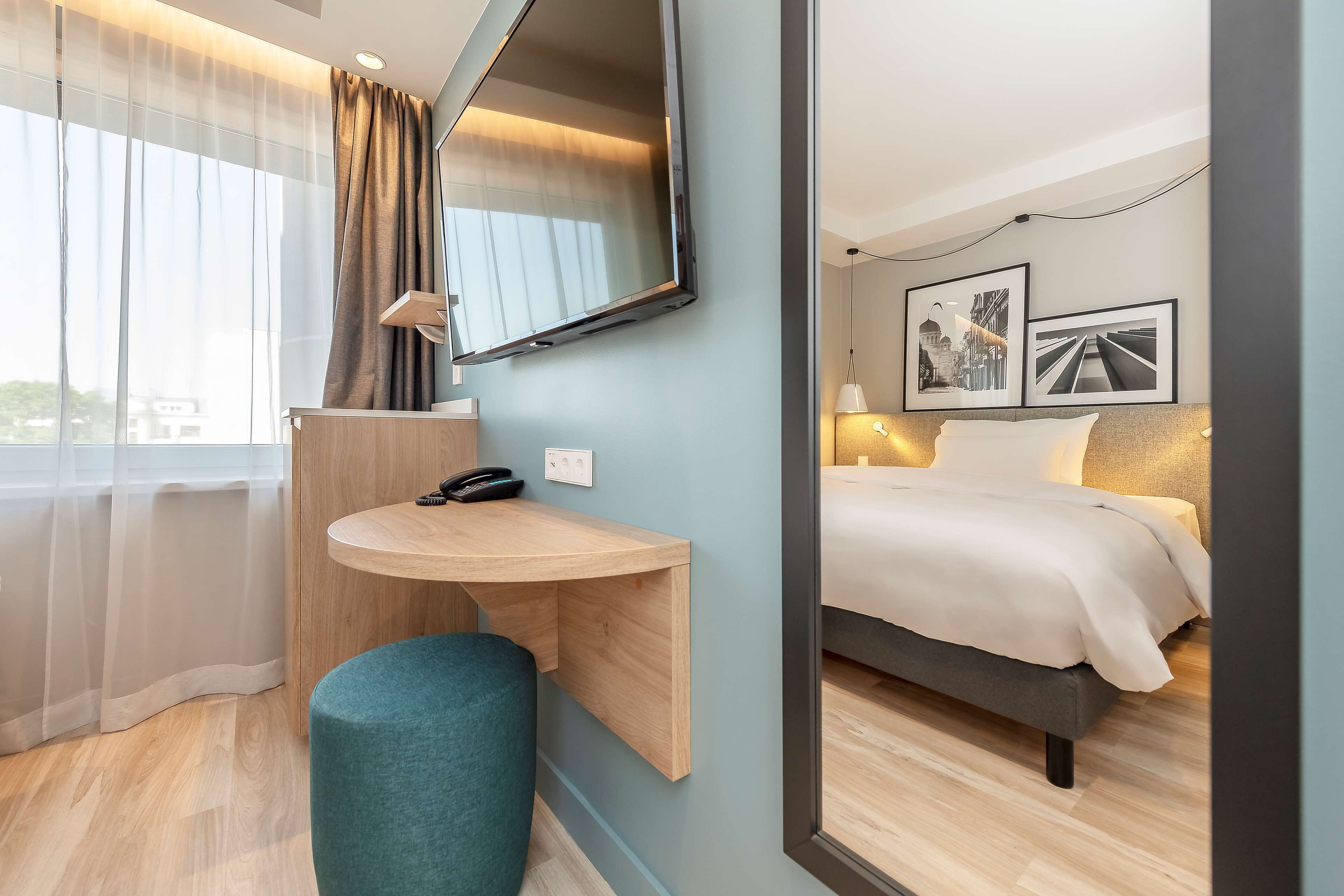 Habitación de hotel moderna con un escritorio de madera, un pouf redondo, una cama grande y un televisor montado en la pared.