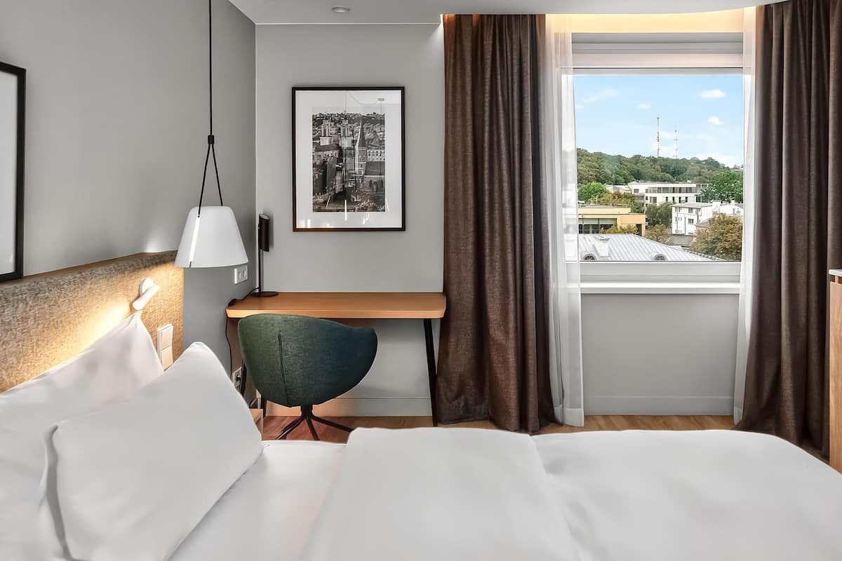 A hotel room with a bed, a desk, a chair, a lamp, a framed picture, and a window with curtains.