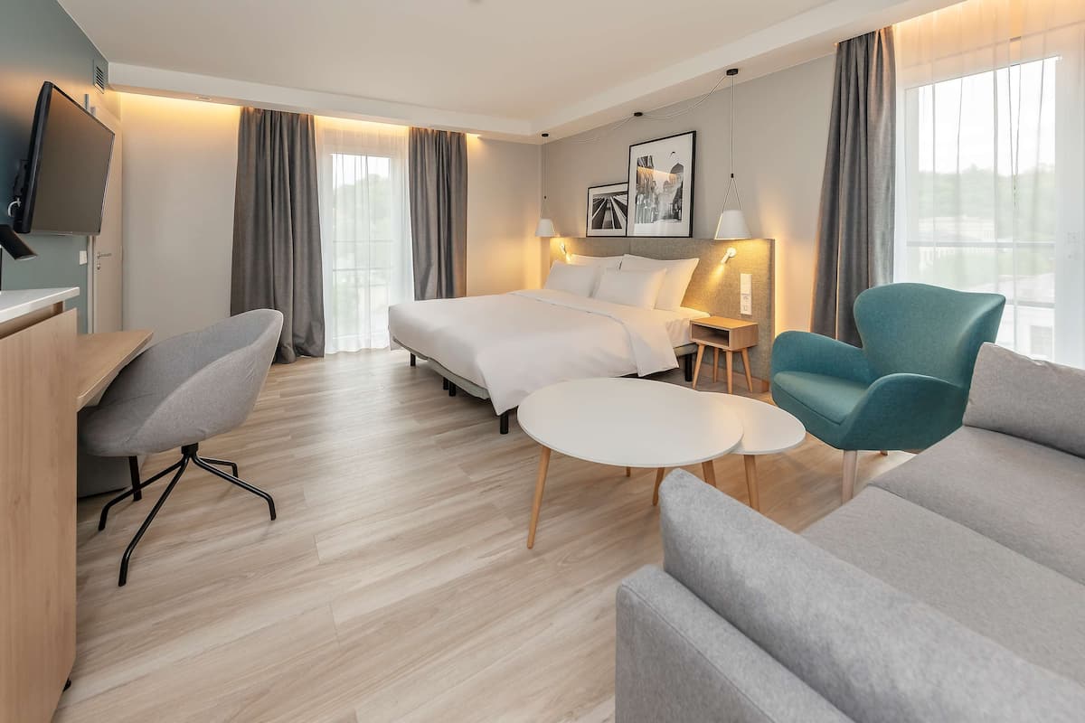 A modern hotel room with a bed, a desk, a chair, a sofa, and a small table.