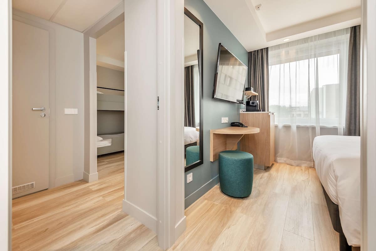 A hotel room with a bed, a desk, a mirror, a window with curtains, and a door.