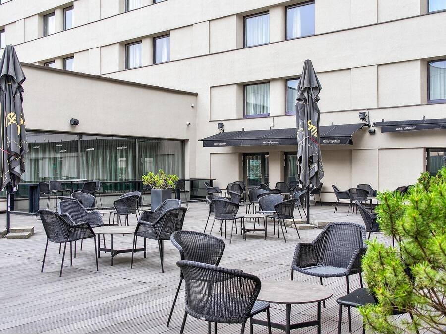 An outdoor patio with wicker chairs and tables, umbrellas, and a building in the background.