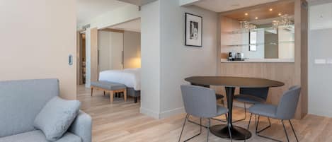Una habitación de hotel moderna con un sofá gris, una mesa redonda con sillas, un espejo grande y un cuadro en la pared.