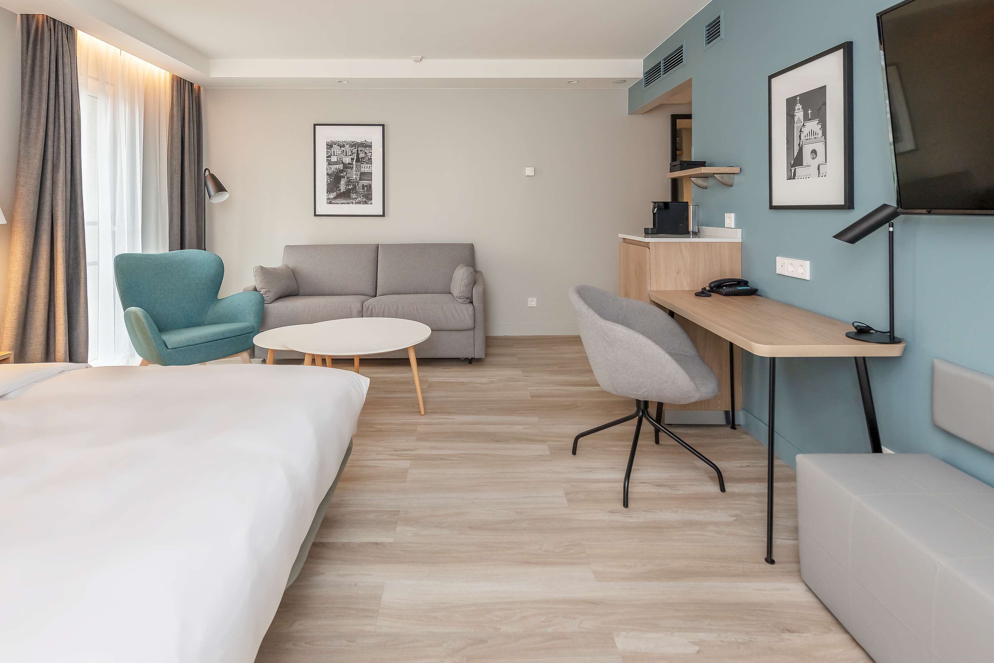 A hotel room with a bed, a sofa, a desk, and a chair. There are two framed pictures on the wall.