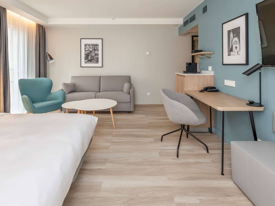 A hotel room with a bed, a sofa, a desk, and a chair. There are two framed pictures on the wall.