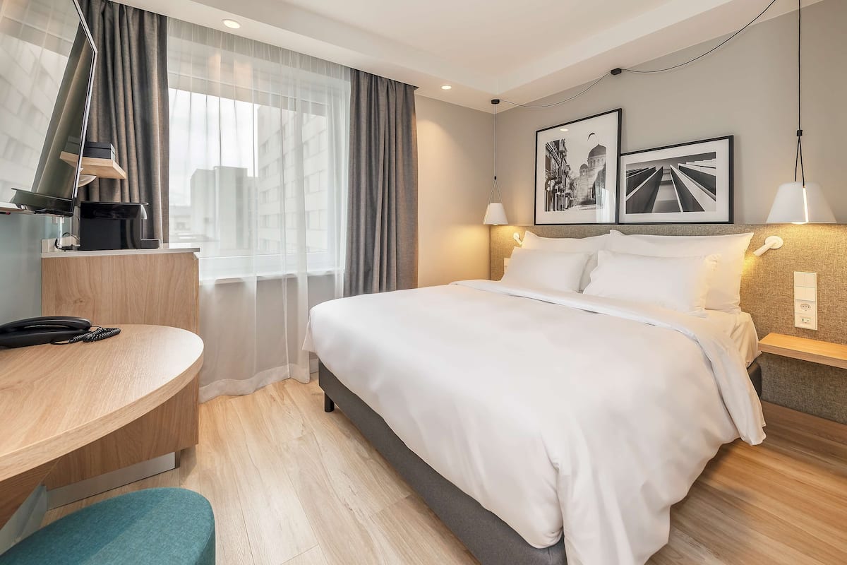 A hotel room with a bed, bedside table, desk, and a large window with curtains.