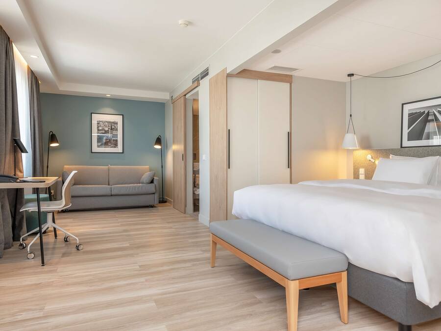 A modern hotel room with a large bed, a desk, a chair, and a sofa.