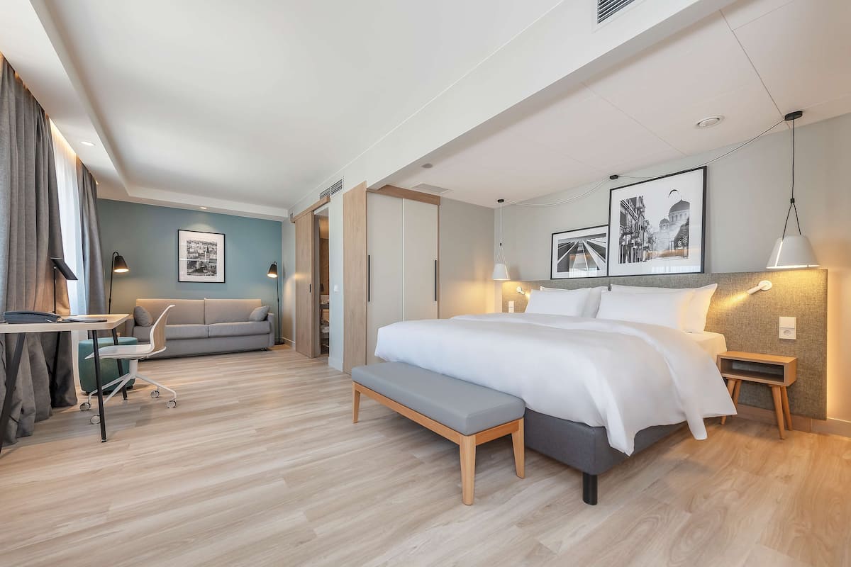 A modern hotel room with a bed, bedside tables, a desk, a chair, and a bench.