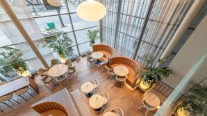 A spacious indoor area with multiple seating arrangements, including round tables and cushioned seating, surrounded by large windows offering views of the street outside. - Radisson Hotel Kaunas (Kaunas)