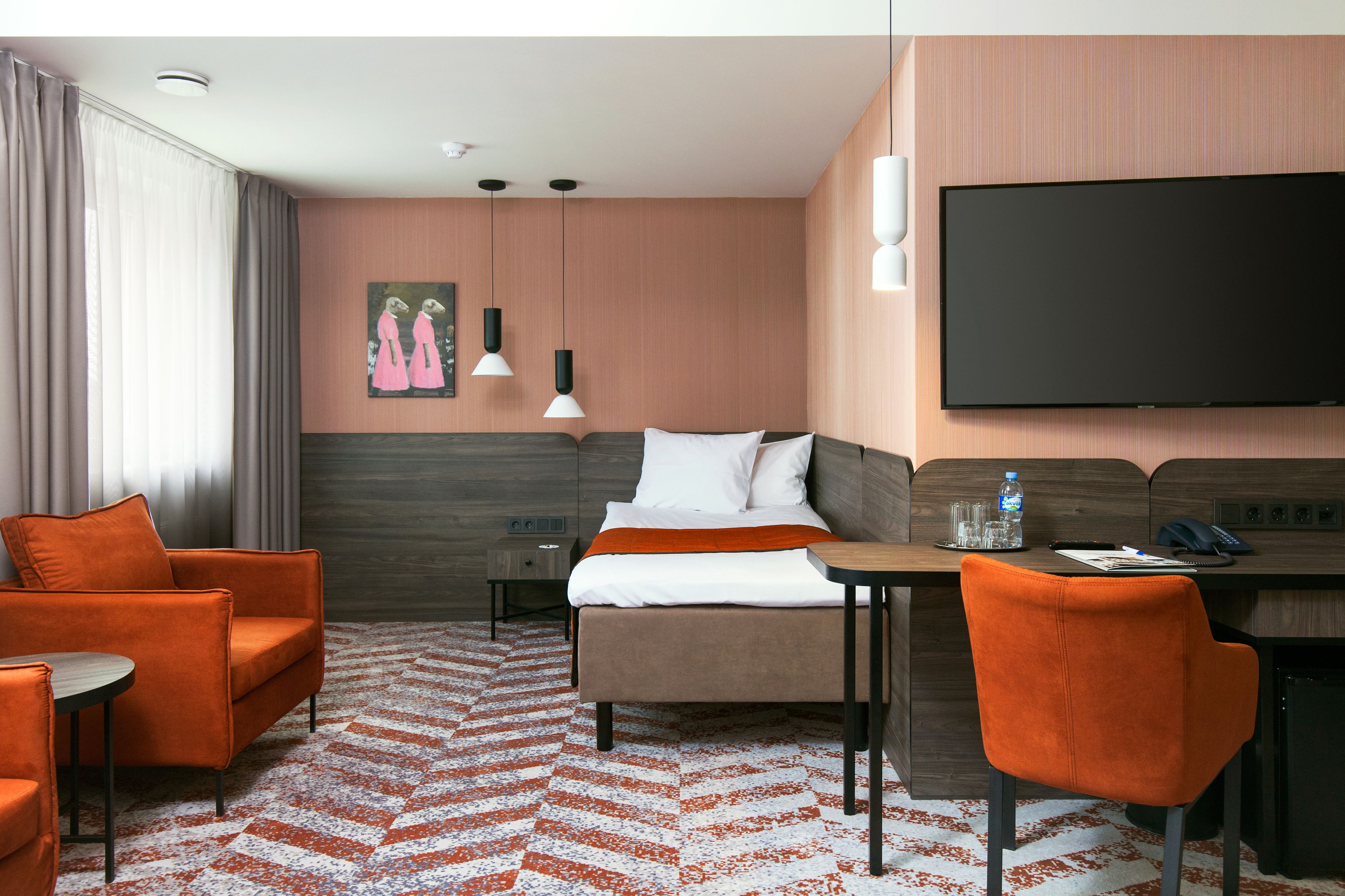 A hotel room with a bed, two orange armchairs, a small table, a desk, a flat-screen TV, and a painting on the wall.