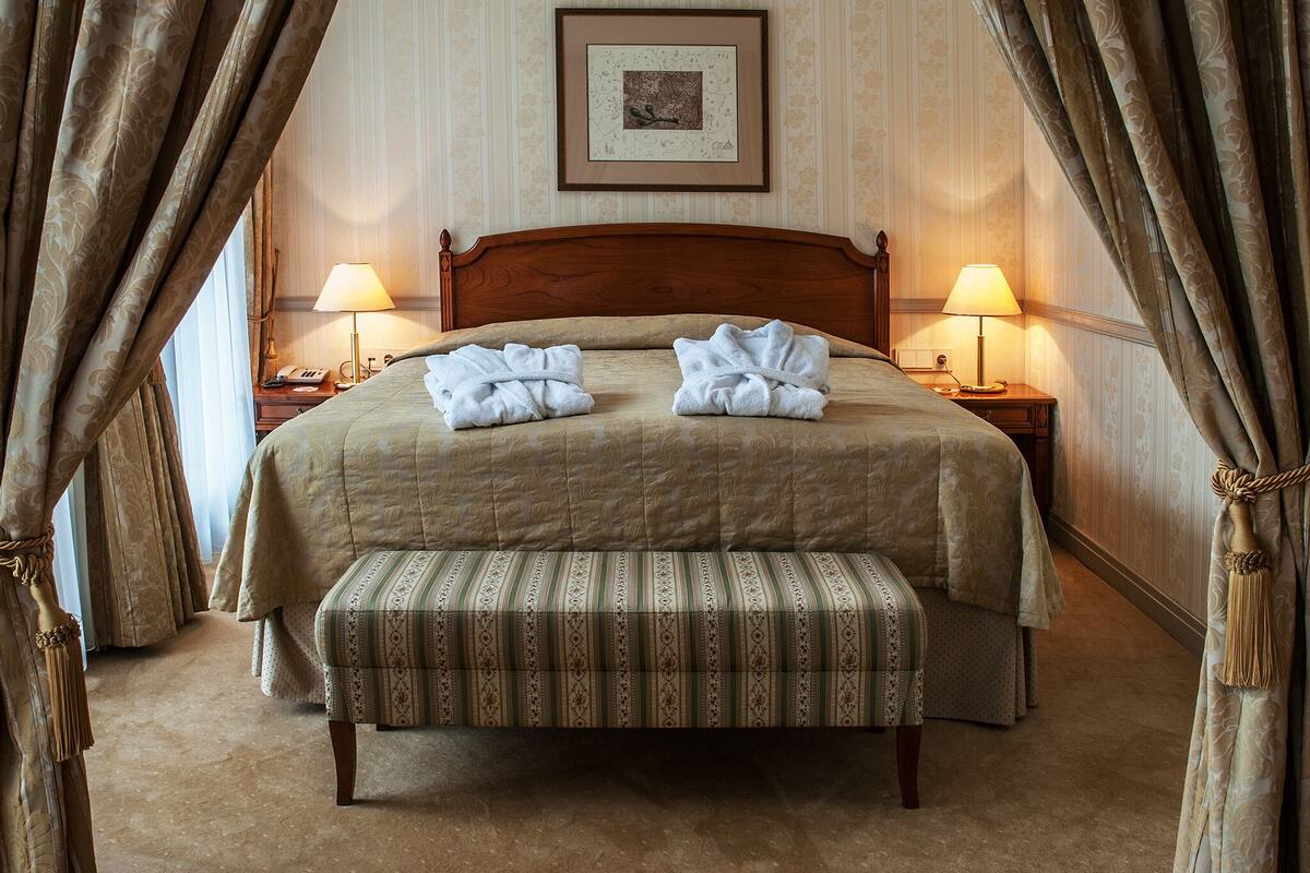 Una cama bien hecha con cabecera de madera, dos lámparas de noche, un cuadro enmarcado en la pared y un banco a rayas frente a la cama.