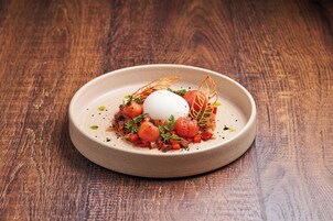 Un plato de cerámica redondo con un postre blanco, decorado con hierbas y pequeñas verduras, sobre una mesa de madera.