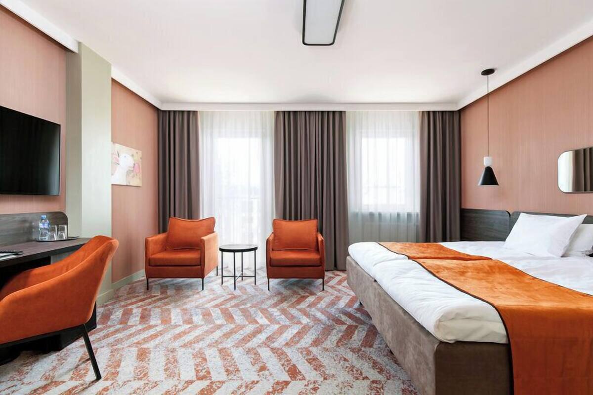 Una habitación de hotel con una cama, dos sillones naranjas, una mesita, un televisor y un ventanal con cortinas.