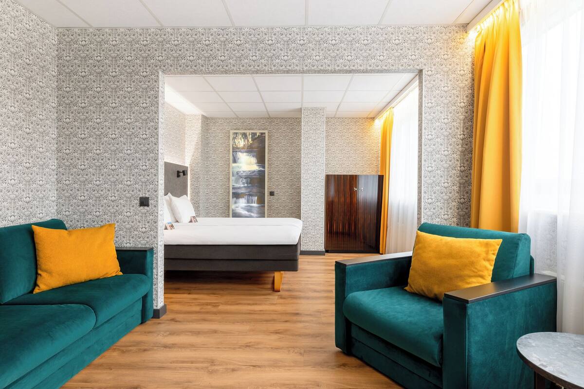 Ein Hotelzimmer mit einem Bett, zwei grüne Sofas mit gelben Kissen, einem Holzboden und Wandtapete mit Muster.