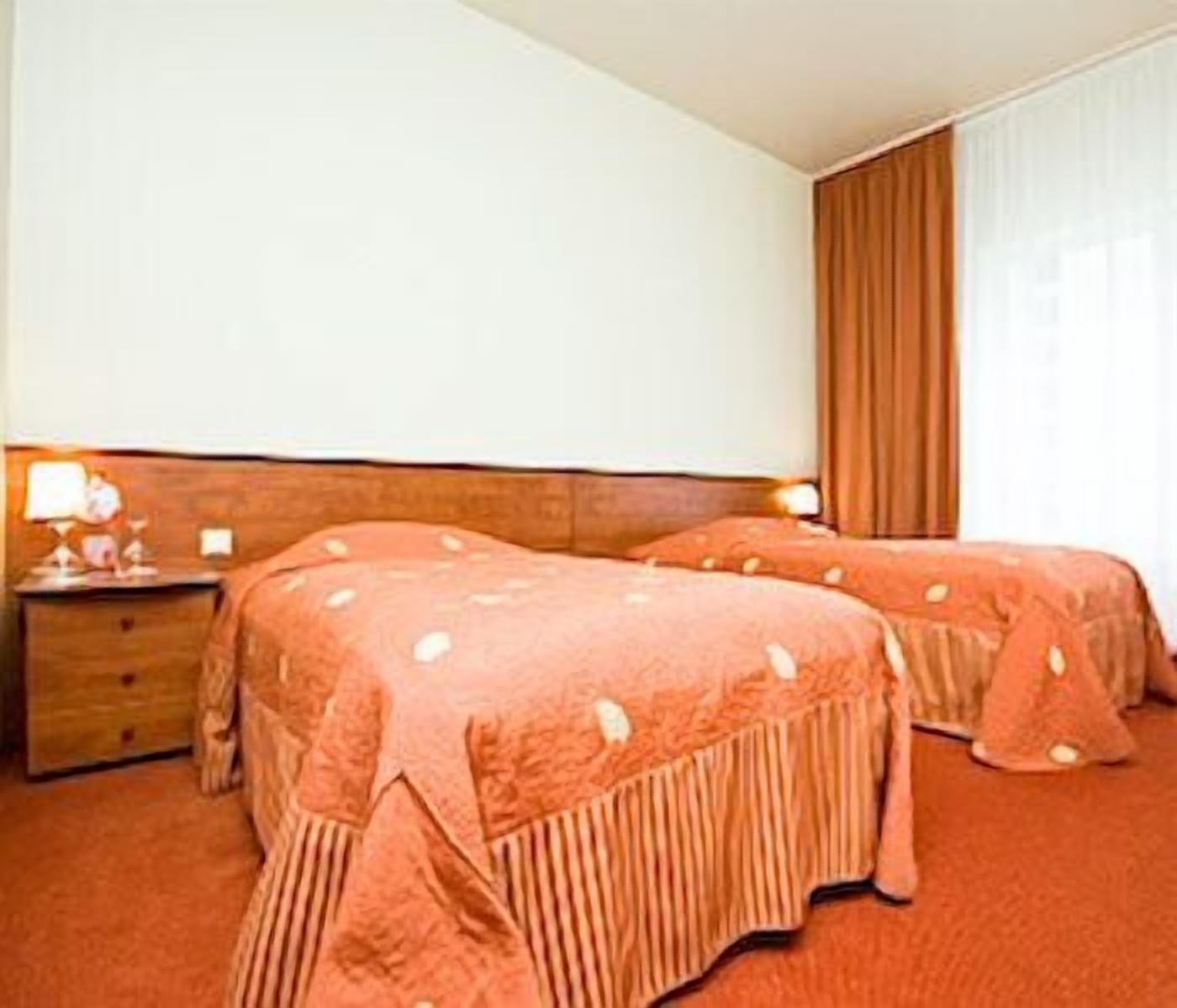 A hotel room with two beds, a wooden headboard, a nightstand with a lamp, and a window with curtains.