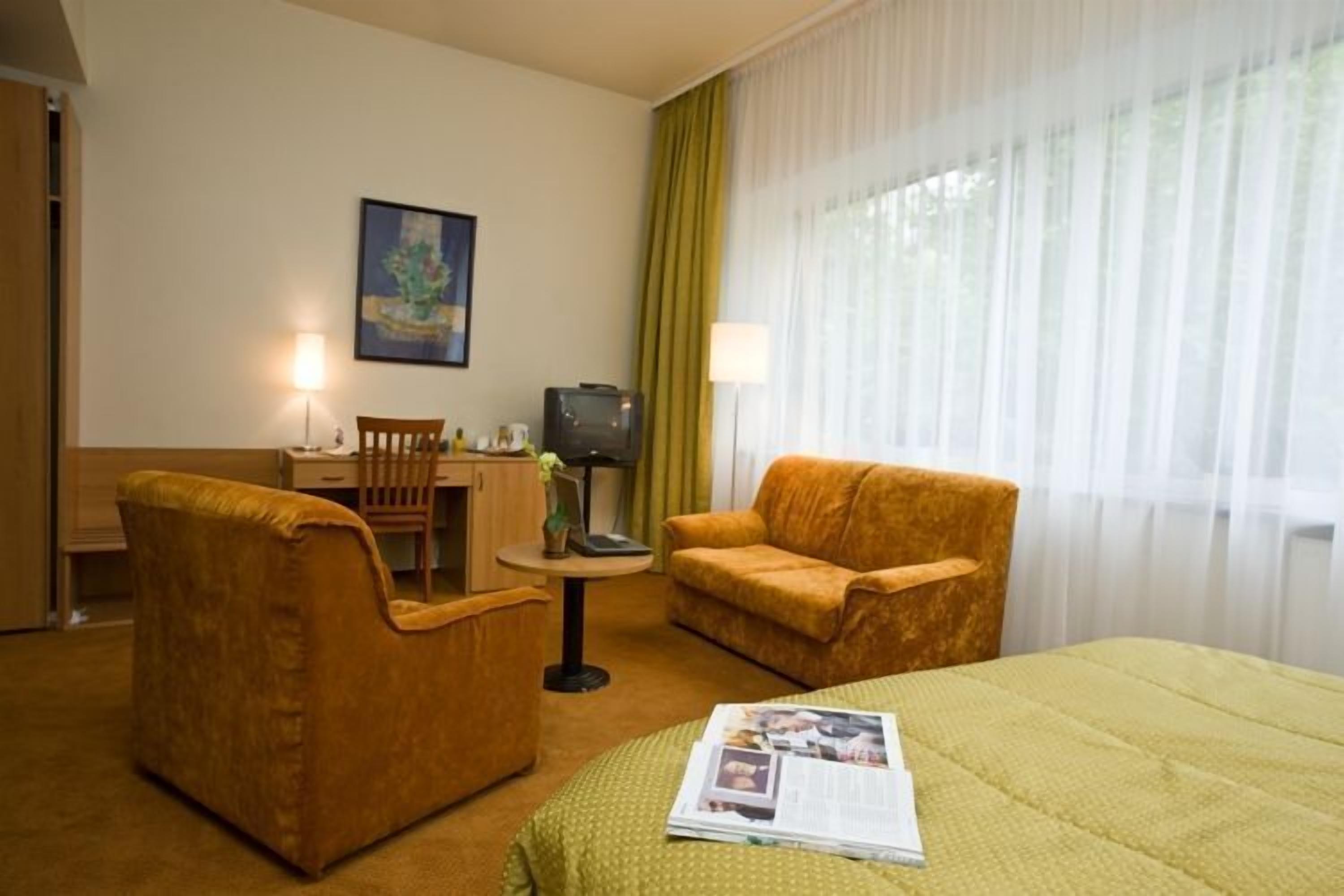 A hotel room with a bed, two armchairs, a small table with a lamp, a television, and a painting on the wall.