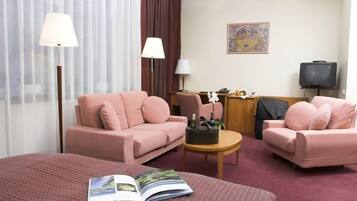 Et hotelværelse med to lyserøde sofaer, en seng med et rødt mønstret sengetæppe, et rundt bord med en lampe, et fjernsyn og et billede på væggen.