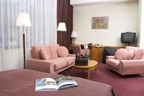A hotel room with two pink sofas, a bed with a red patterned bedspread, a small round table with a lamp, a television, and a painting on the wall. - Best Baltic Hotel Kaunas (Kaunas)