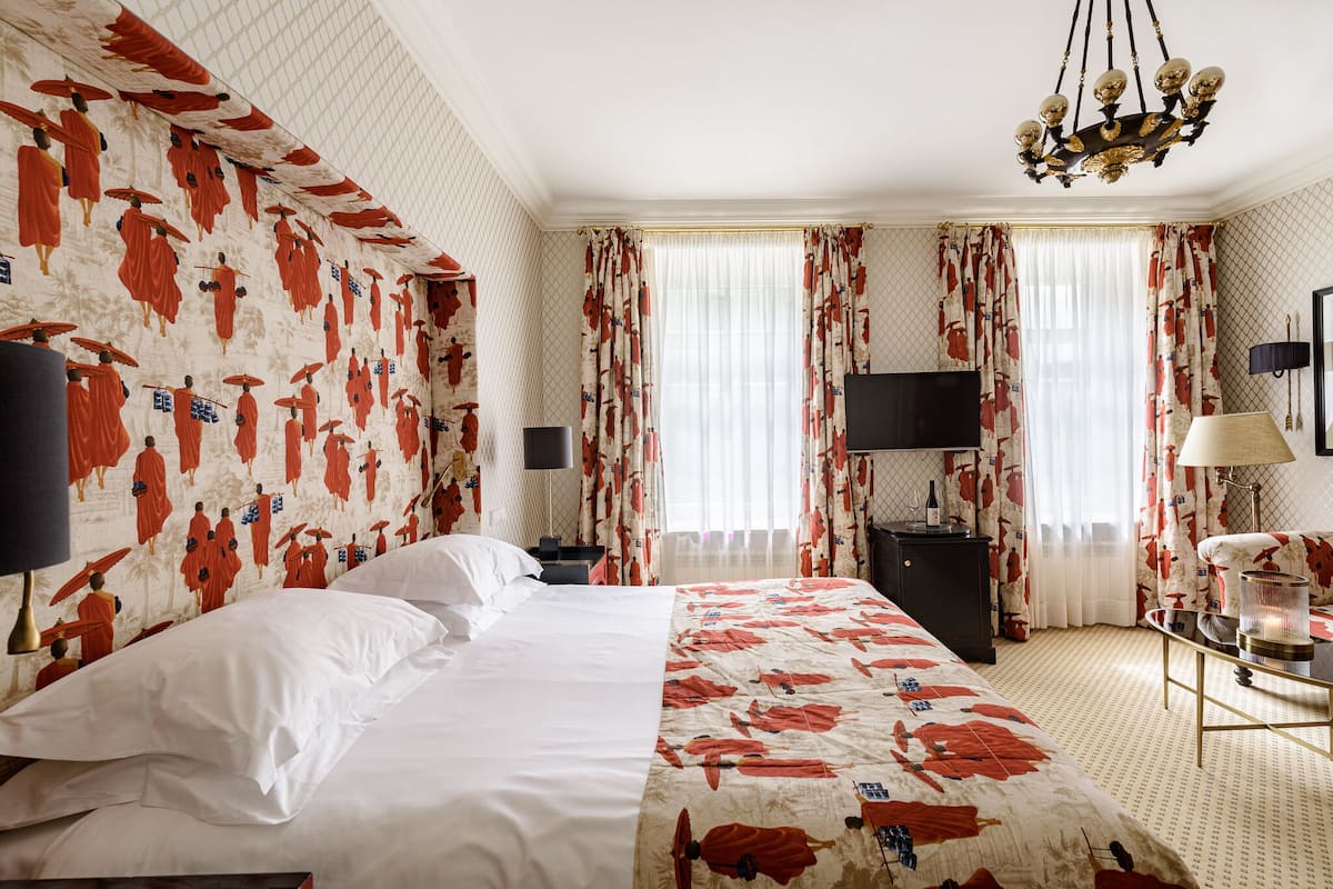 Una habitación de hotel con una cama grande, un televisor de pantalla plana, un escritorio con una lámpara y cortinas con estampado rojo y blanco.