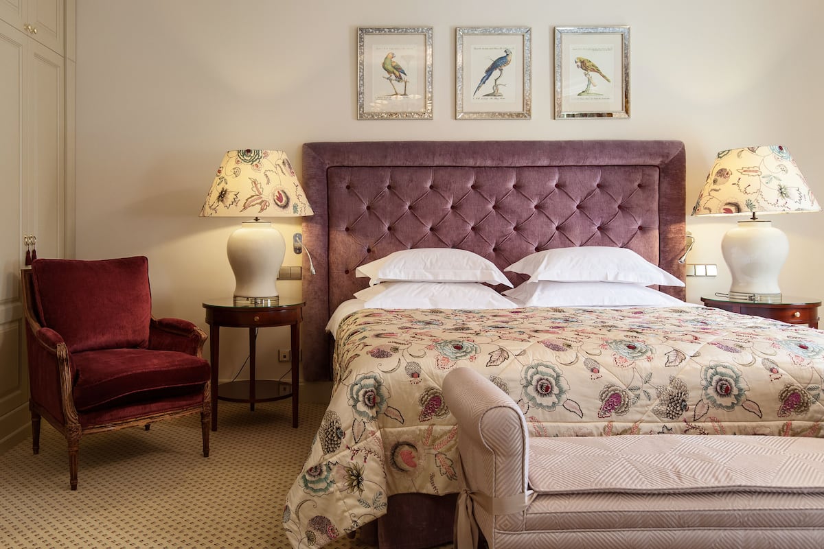 Un dormitorio con cabecera capitonada, colcha con estampado floral, dos cuadros enmarcados con pájaros, un sillón rojo y dos mesitas de noche con lámparas.