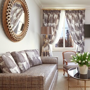 A room with a patterned sofa, a round table with a vase of flowers, a lamp, and curtains with a historical print. - NARUTIS hotel (Vilnius)