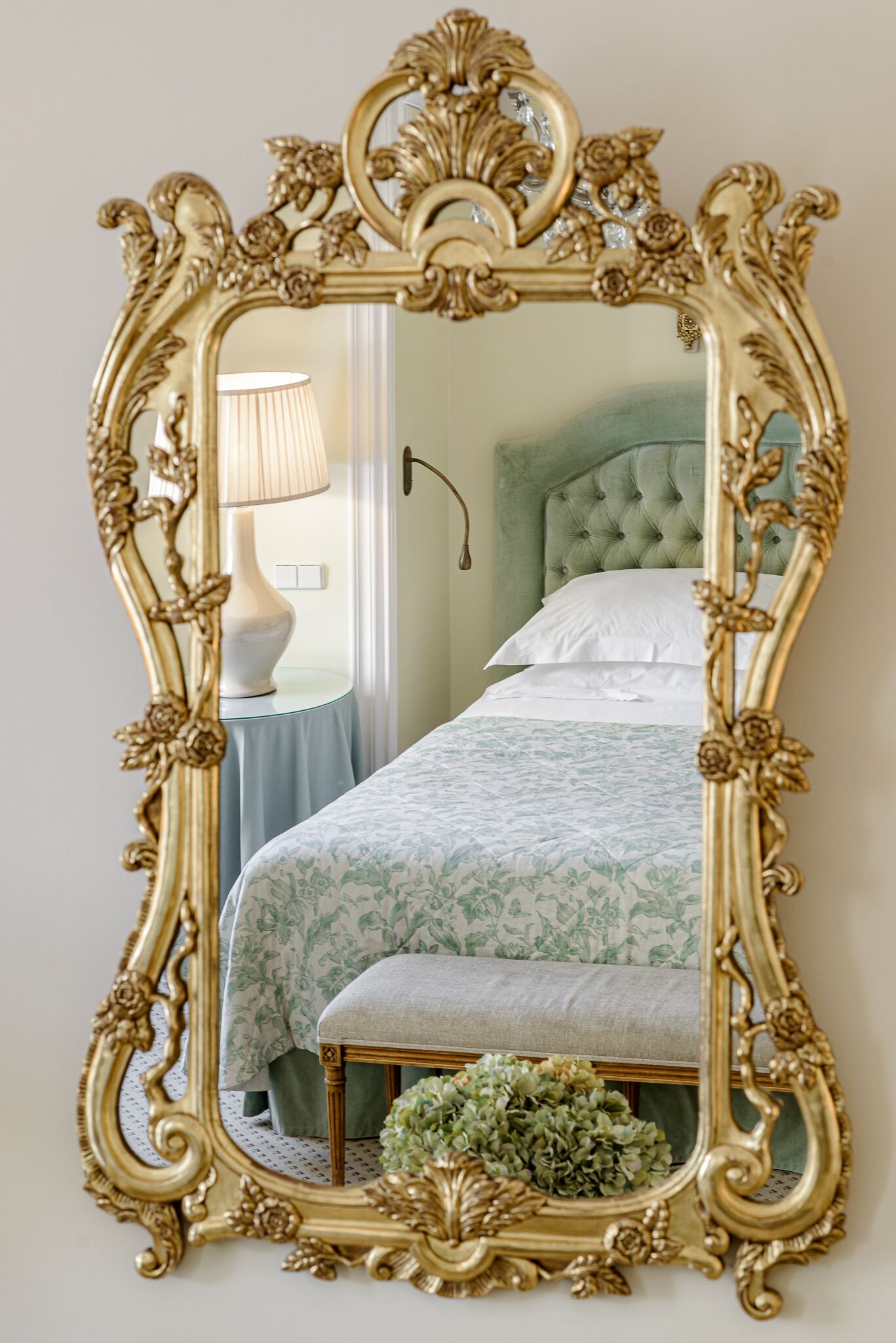 Une chambre à coucher avec un grand miroir décoratif, une tête de lit recouverte d’un tissu vert, une table de chevet avec une lampe et une couvre-lit à motifs floraux.