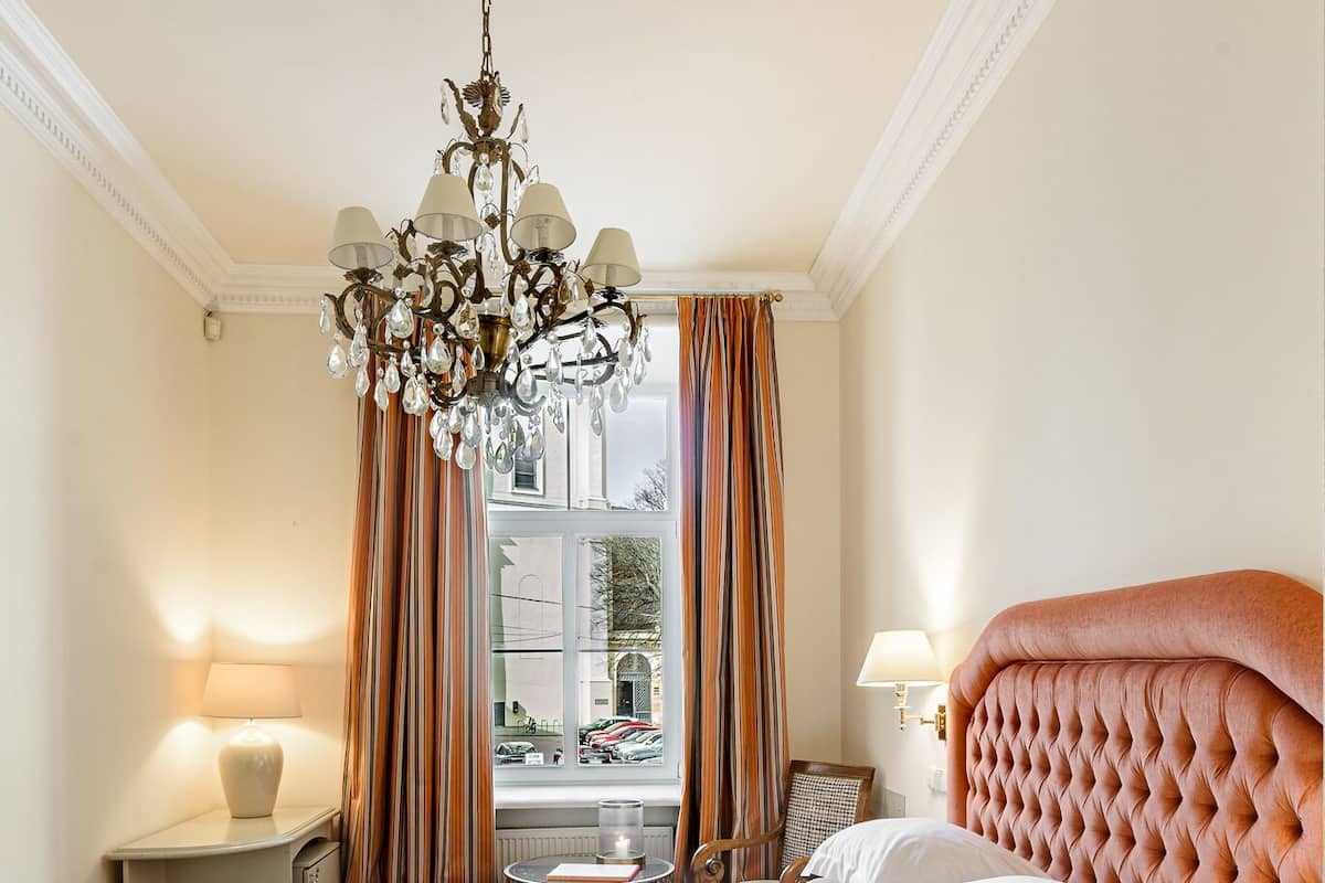 Habitación de hotel con una cama grande, una araña, una ventana con cortinas y vistas a la calle.