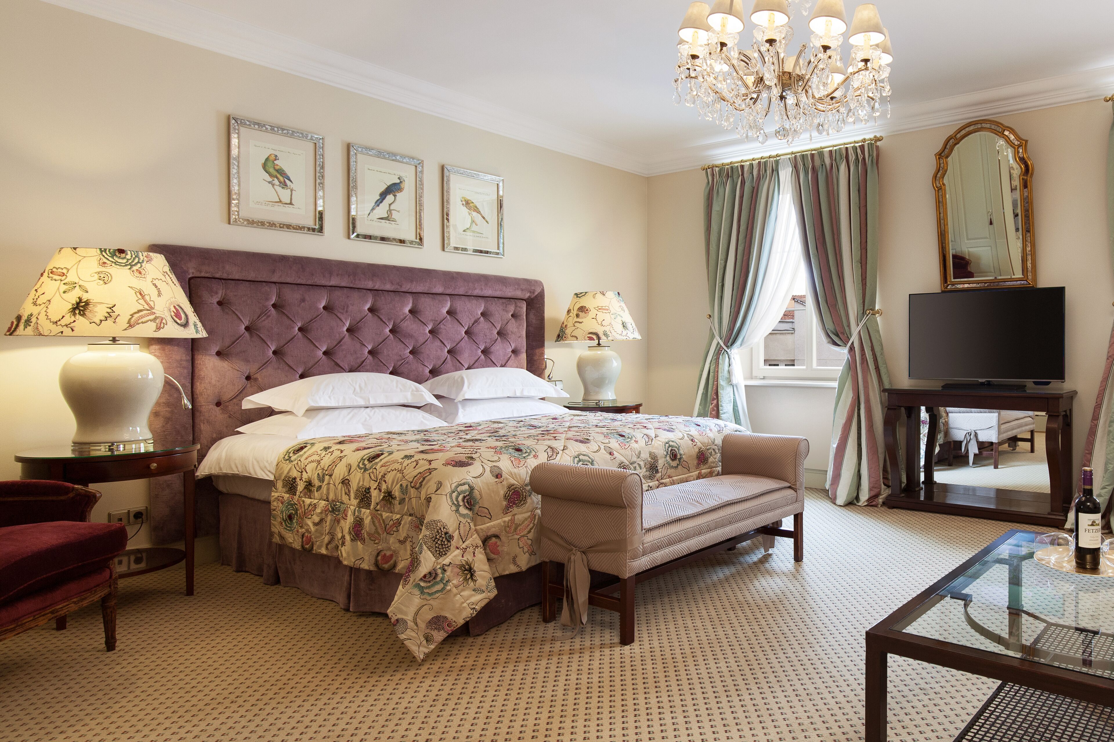 A hotel room with a large bed, two bedside lamps, a television, a chair, and a chandelier.