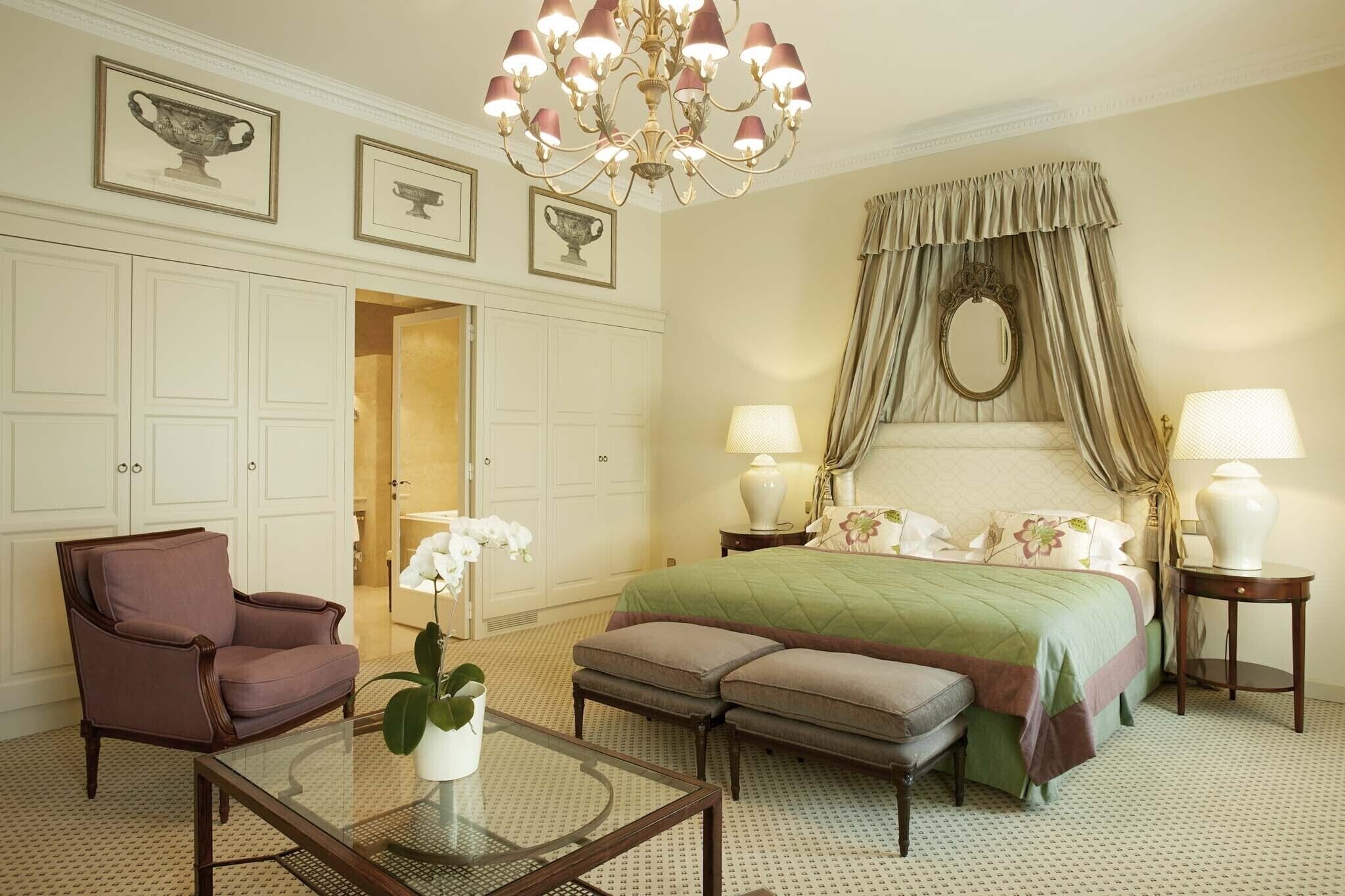 A hotel room with a large bed, a sitting area with a chair and ottoman, a glass coffee table, and a chandelier.